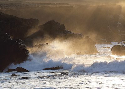 Waves crashing against rocky cliffs, illuminated by sunlight, creating a dramatic coastal scene.