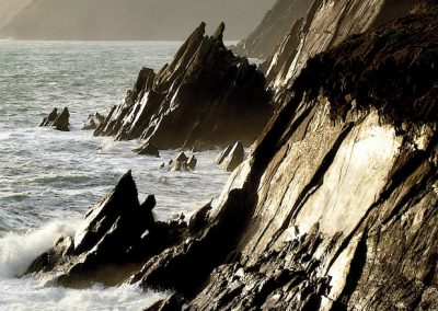 Jagged coastal rocks with crashing waves. Mountains in the distance.
