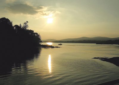 Sunset over a tranquil lake with silhouetted trees and distant mountains. Gallery Two.