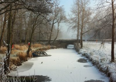 Frozen river scene in winter with a boat and bare trees under a bridge. From Gallery Two.