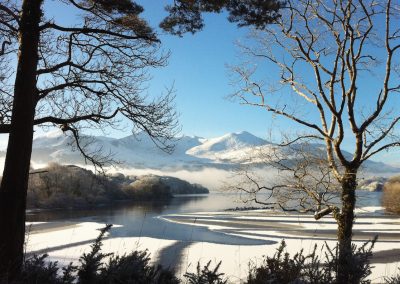 Winter landscape with snow-covered mountains, lake, and trees under a clear blue sky.